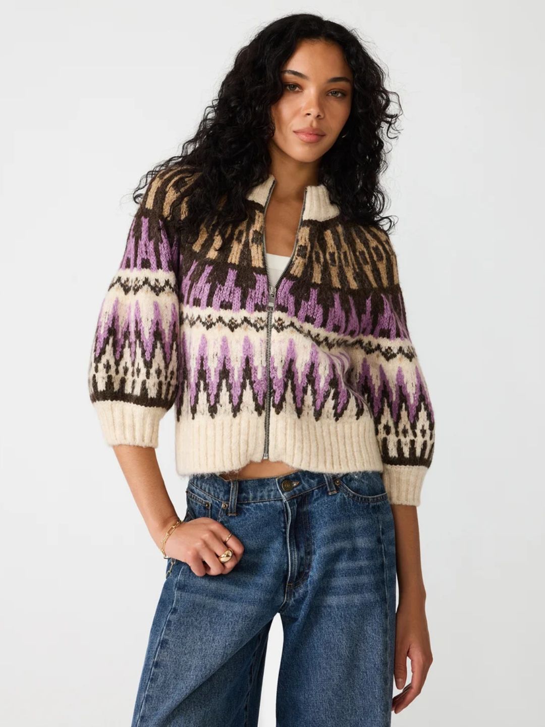 A woman modeling a cropped knit zip-front cardigan with a Fair Isle pattern in cream, brown, and purple, paired with blue jeans against a plain white studio background.