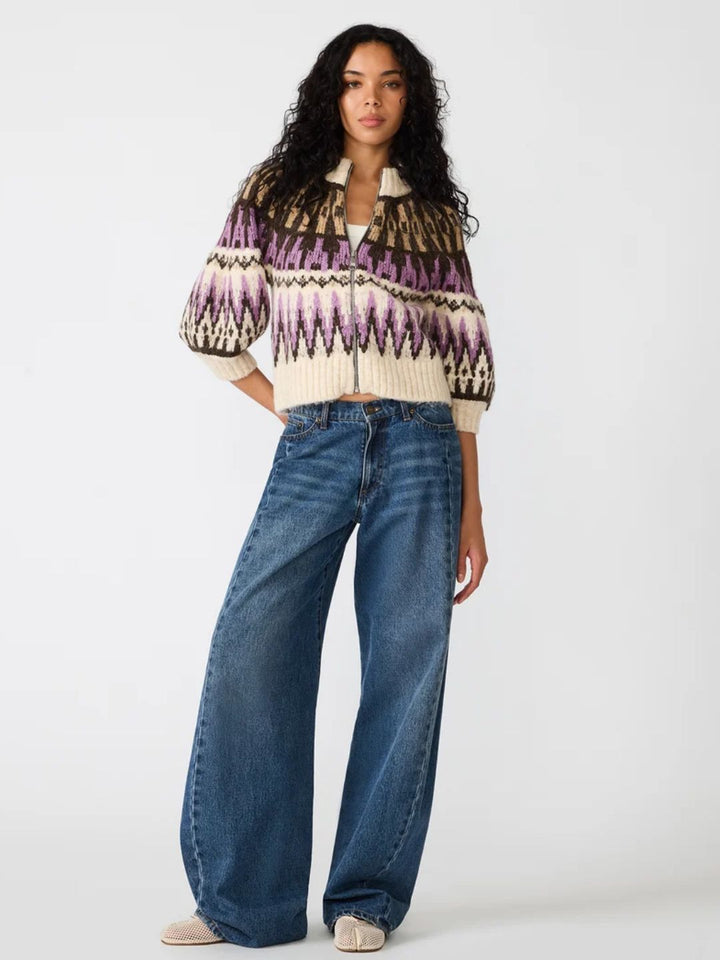 A model wearing a cream knit cardigan with purple, black, and brown geometric patterns, paired with wide-leg blue jeans and beige shoes, against a plain white studio background.