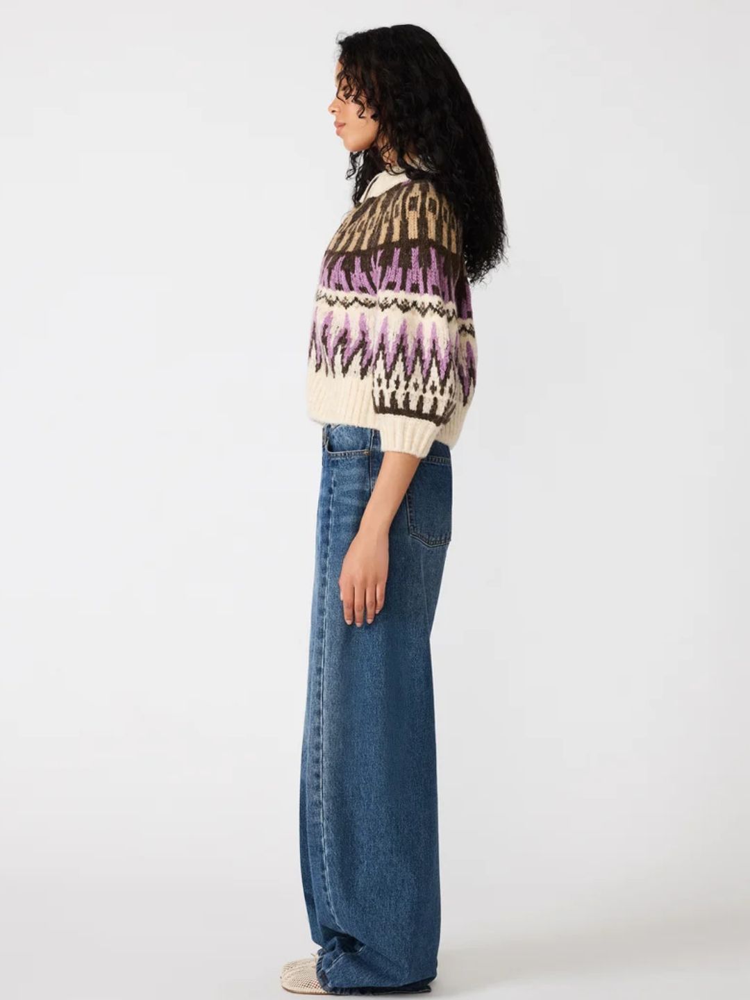 A woman stands in profile wearing a cream knit sweater with a geometric Fair Isle pattern in purple, brown, and black, paired with wide-leg blue jeans against a plain white studio background.