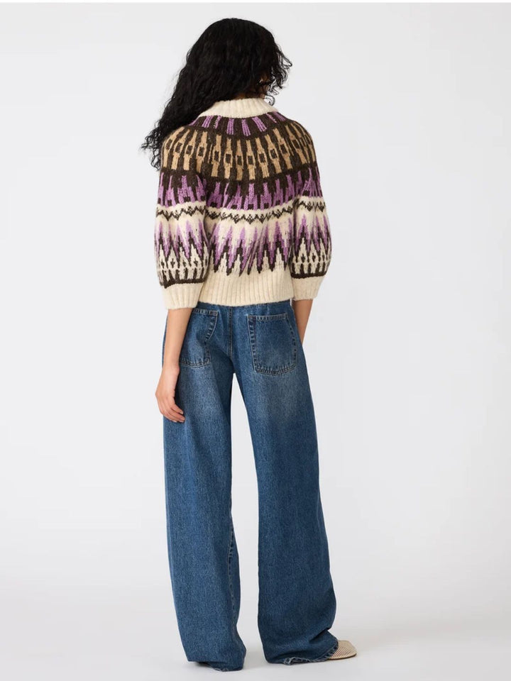 A model shown from the back wearing a cream knit cardigan with a brown, purple, and black geometric pattern, paired with wide-leg blue jeans, against a plain white studio background.