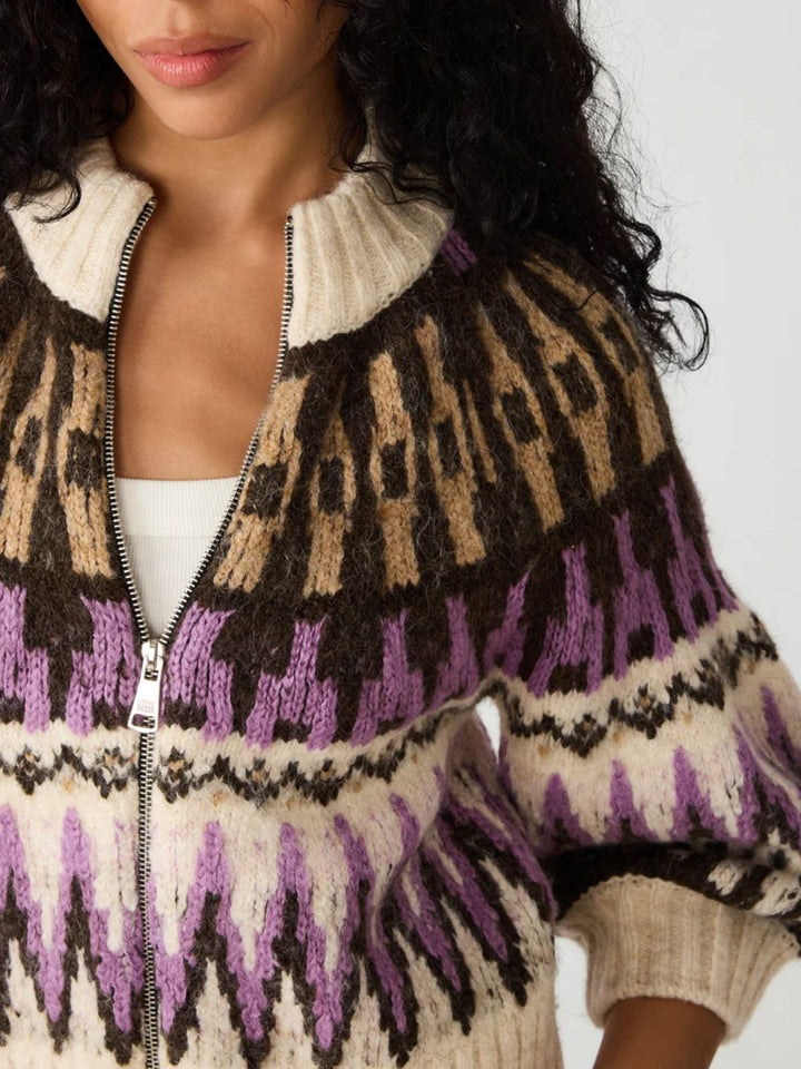 A close-up of a woman wearing a fuzzy knit zip-front sweater with a cream ribbed collar and cuffs, brown and tan geometric bands, and purple and cream zigzag patterns.