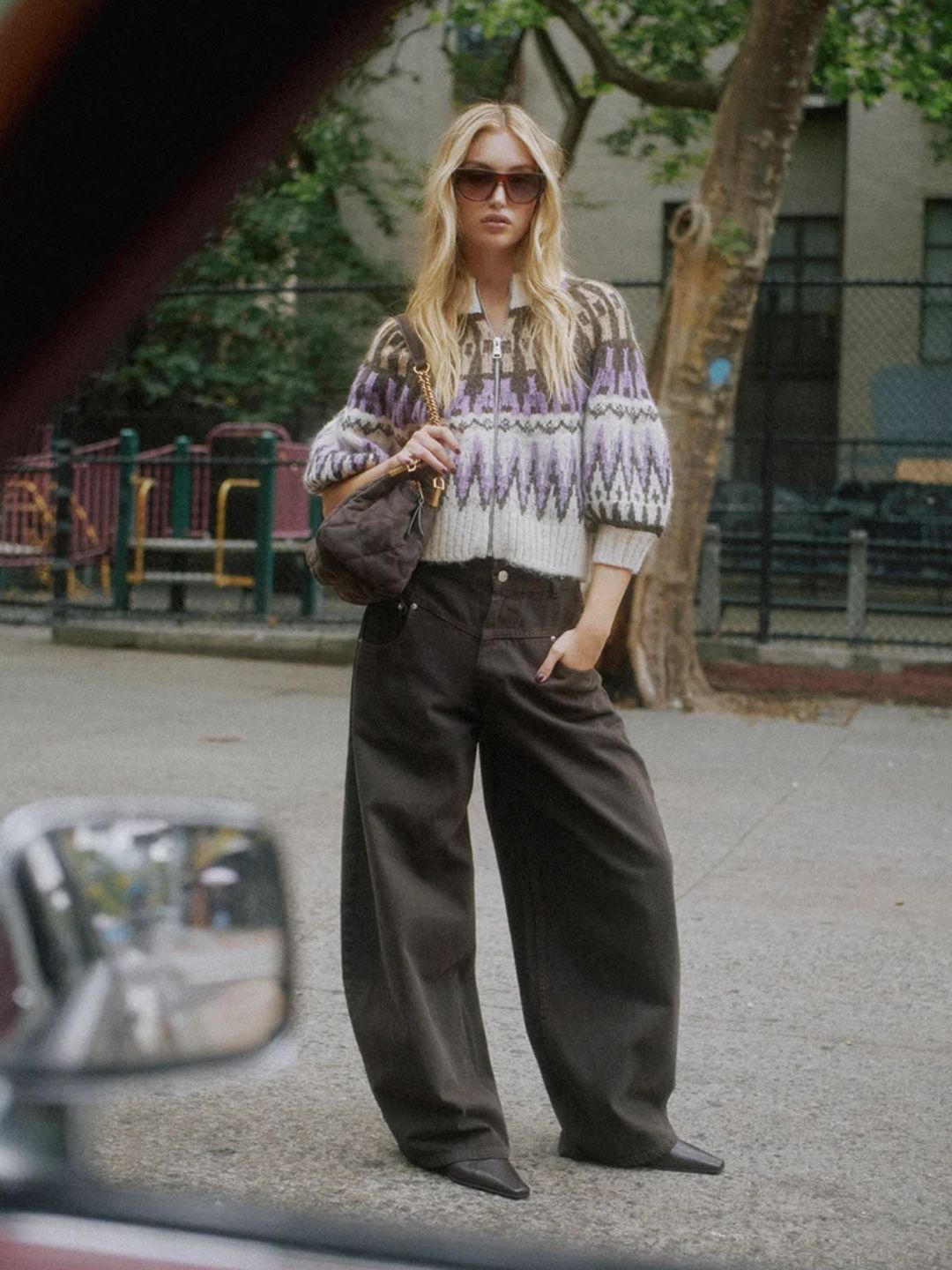 A woman stands outdoors on a street wearing a cream knit sweater with purple and gray geometric patterns, dark wide-leg pants, and sunglasses.