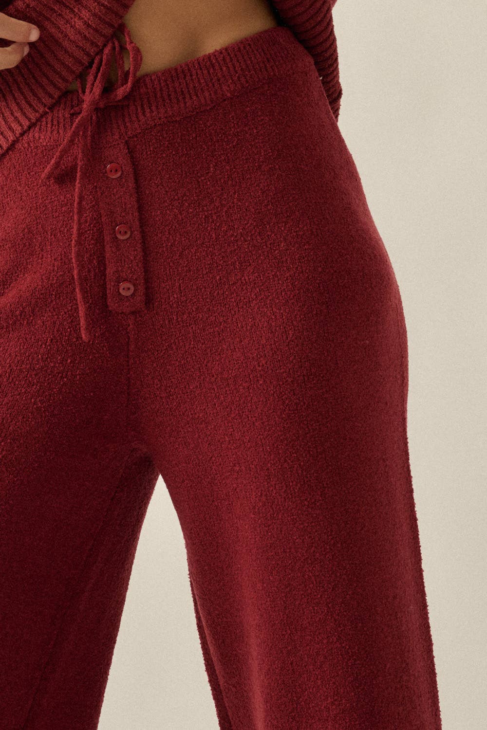 Close-up of a person wearing ribbed knit lounge pants in a deep burgundy red, featuring a drawstring waistband and front button detail.