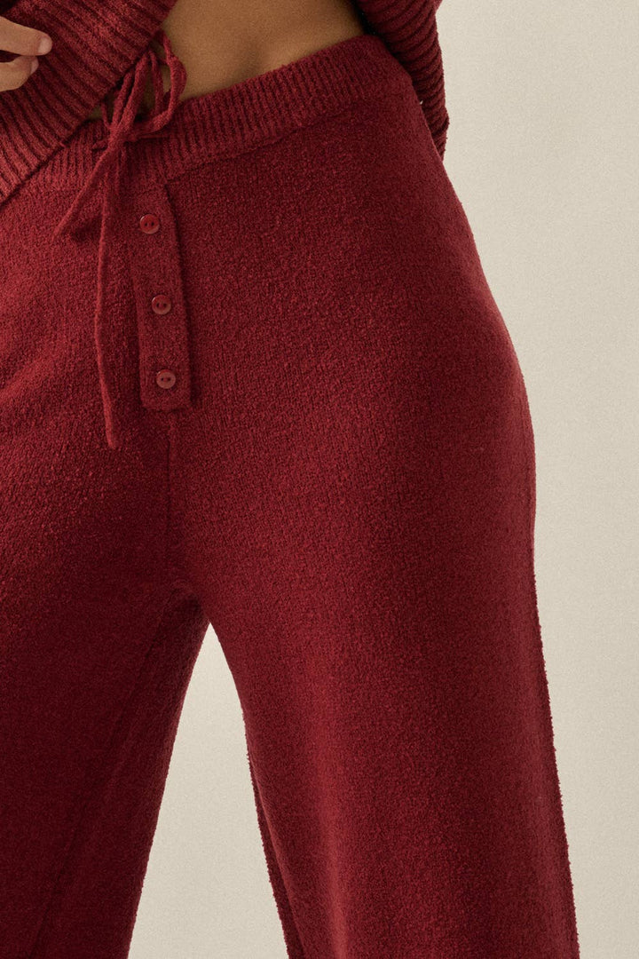 Close-up of a person wearing ribbed knit lounge pants in a deep burgundy red, featuring a drawstring waistband and front button detail.