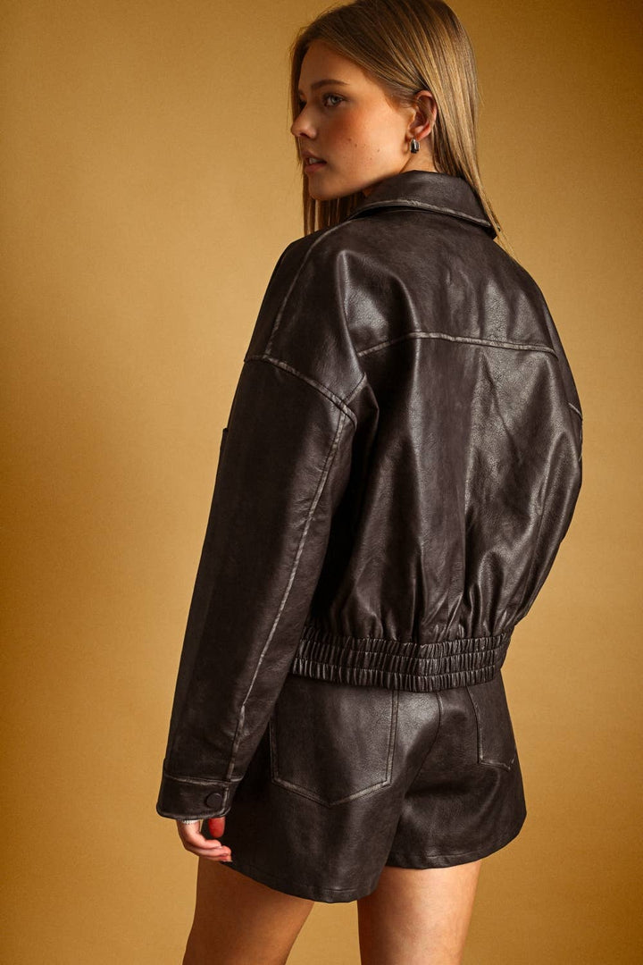 A woman wearing a dark brown faux leather bomber jacket and matching leather shorts, shown from a side-back angle against a warm tan studio background.