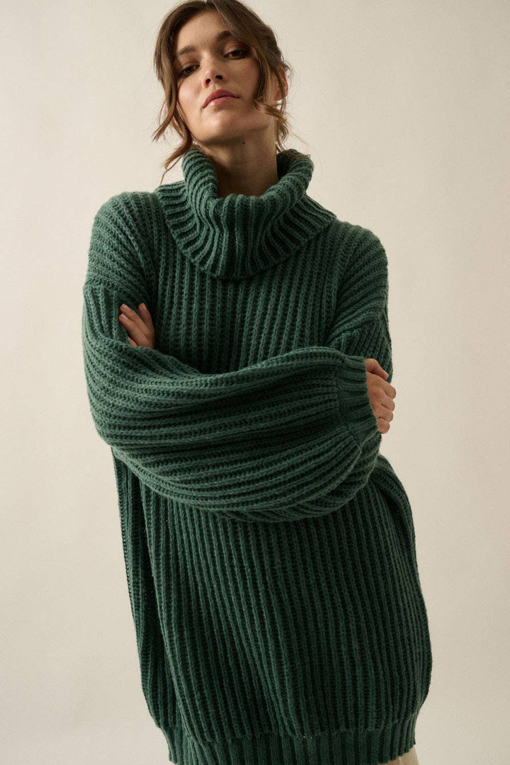 A woman wearing an oversized dark green ribbed knit turtleneck sweater, posing with her arms crossed in front of a plain light beige background.