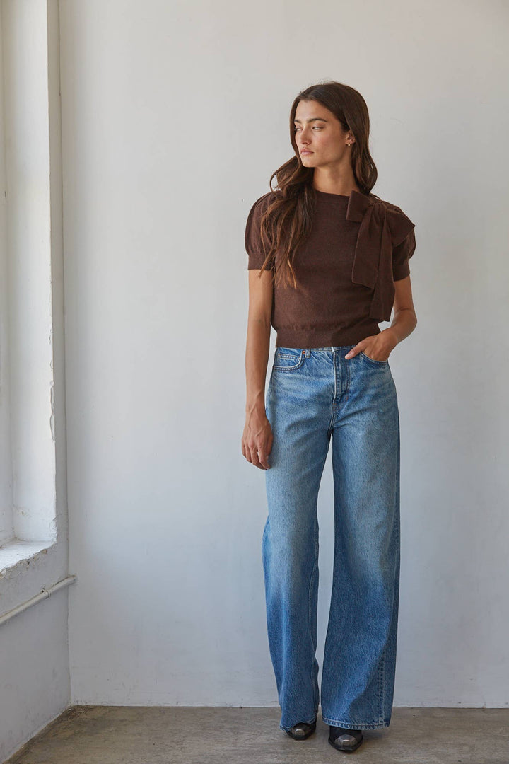 A woman modeling a brown short-sleeve top and wide-leg blue jeans, standing in a minimalist room with a white wall and concrete floor.