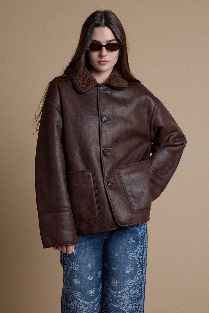 A woman wearing a reversible dark brown leather jacket with a brown sherpa on reverse side, paired with blue paisley-print jeans, against a solid beige studio background.