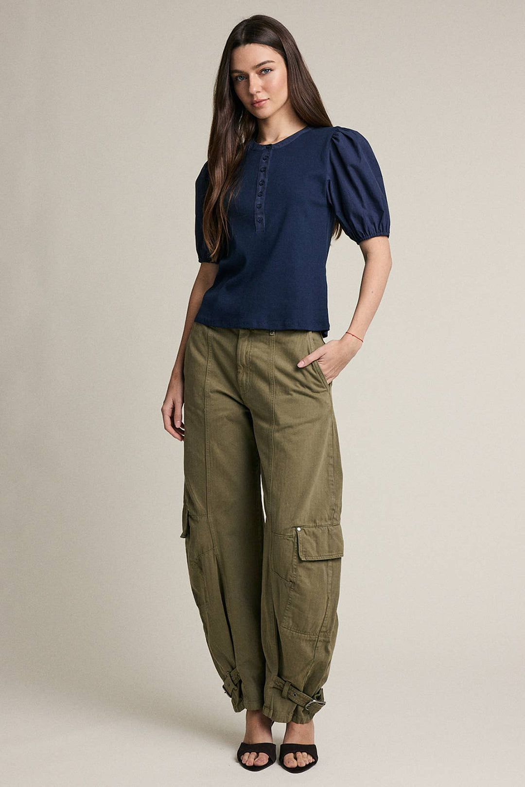 A woman modeling a navy blue short-sleeve top with puff sleeves and olive green cargo pants, standing against a plain beige studio background.