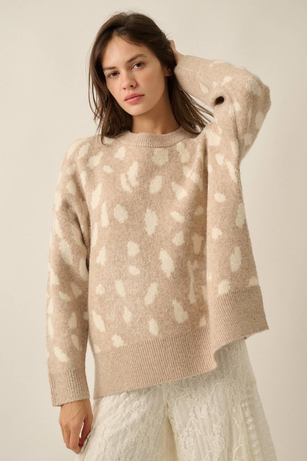 A woman wearing a beige knit sweater with a white abstract spotted pattern, paired with white textured pants, against a plain beige studio background.
