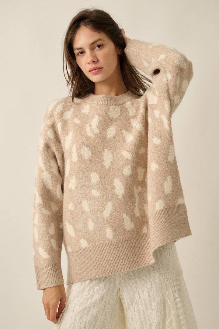 A woman wearing a beige knit sweater with a white abstract spotted pattern, paired with white textured pants, against a plain beige studio background.