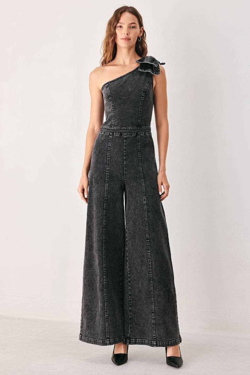 A woman modeling a dark gray washed denim one-shoulder wide-leg jumpsuit with a large bow detail on the shoulder, paired with black heels, against a plain light gray studio background.