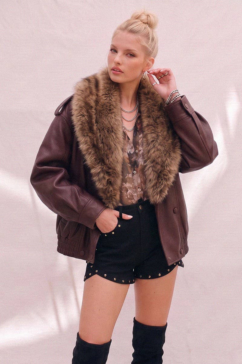 A woman models a burgundy faux-leather jacket with a large brown faux-fur collar, paired with black studded shorts and black over-the-knee boots against a light pink textured backdrop.