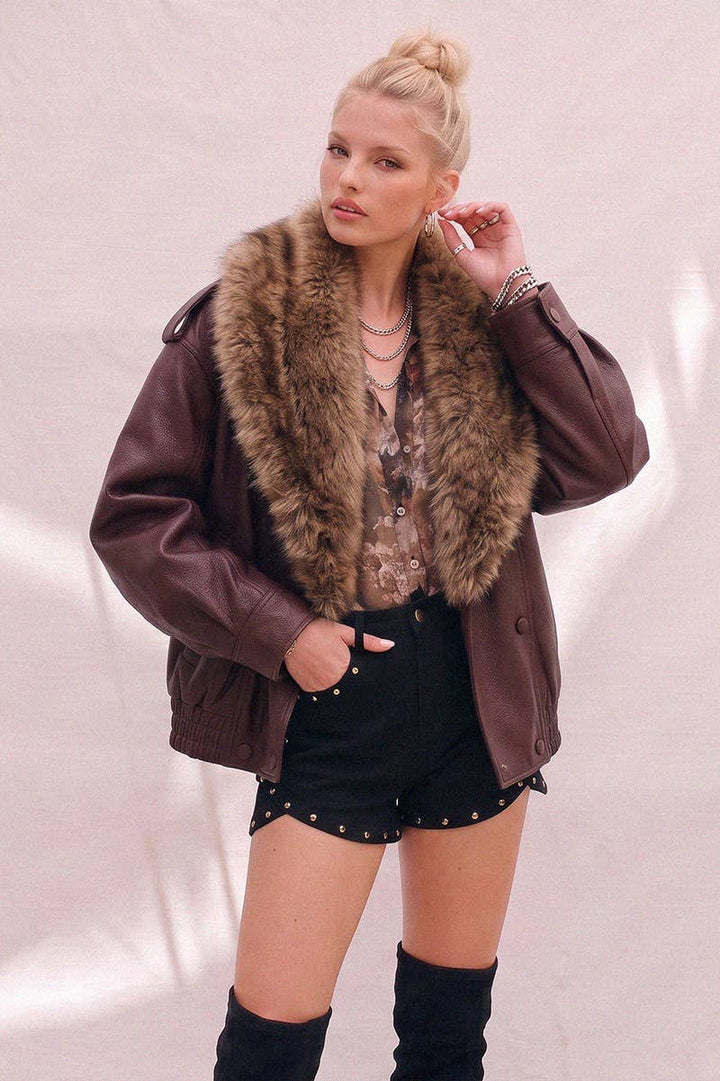 A woman models a burgundy faux-leather jacket with a large brown faux-fur collar, paired with black studded shorts and black over-the-knee boots against a light pink textured backdrop.