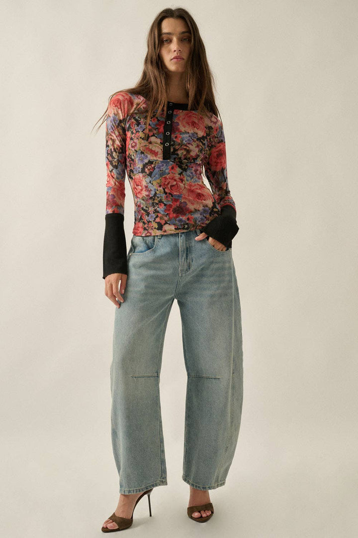 A model wearing a long-sleeve floral print top with black cuffs, paired with wide-leg light-wash jeans and brown heeled sandals, standing against a plain light beige studio background.