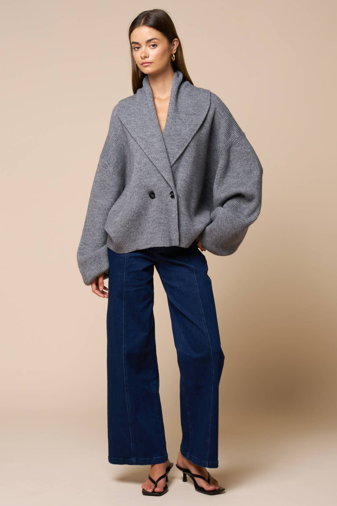 Woman wearing a gray oversized cardigan sweater and blue pants against a beige background