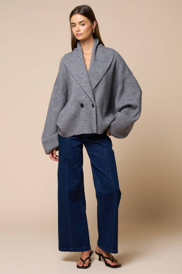 Woman wearing a gray oversized cardigan sweater and blue pants against a beige background