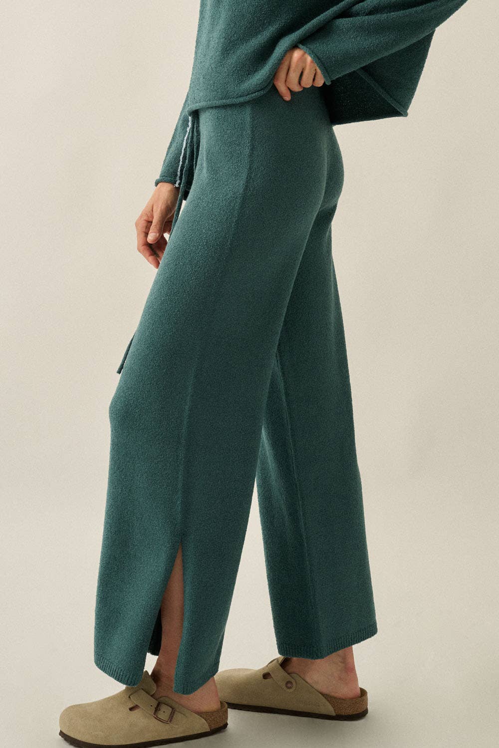 A woman wearing teal wide-leg knit pants with side slits, paired with taupe clogs and a matching teal top, photographed against a plain light beige studio background.