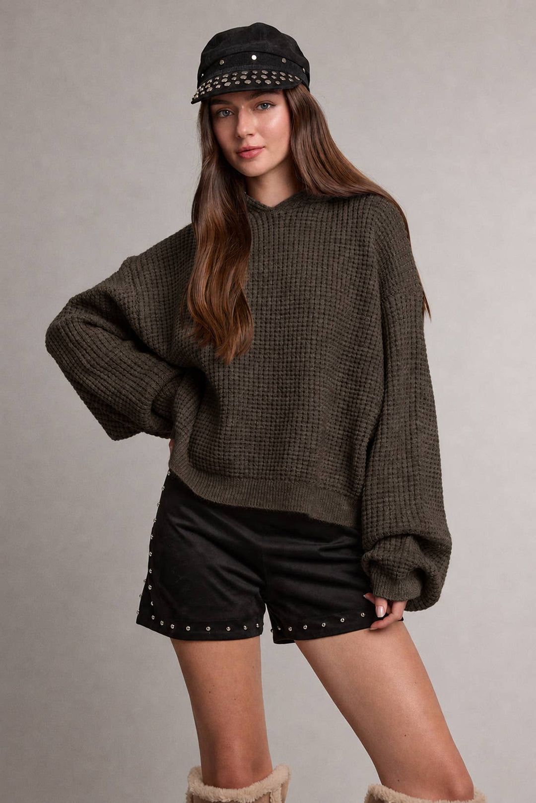 A woman modeling an oversized dark brown waffle-knit hooded sweater, black studded shorts, a black studded cap, and fuzzy beige boots against a neutral gray studio background.