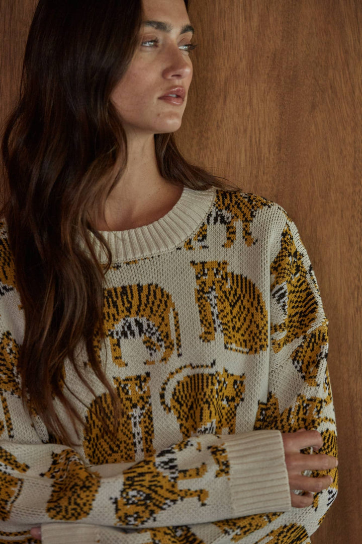 A woman wearing a cream knit sweater with a bold yellow and black tiger print, posed against a warm wood-paneled background.