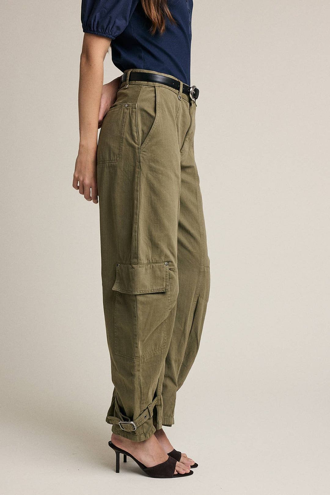 A model is shown from the waist down wearing olive green cargo pants with a black belt, paired with a navy top and black heeled sandals, against a solid light beige background.