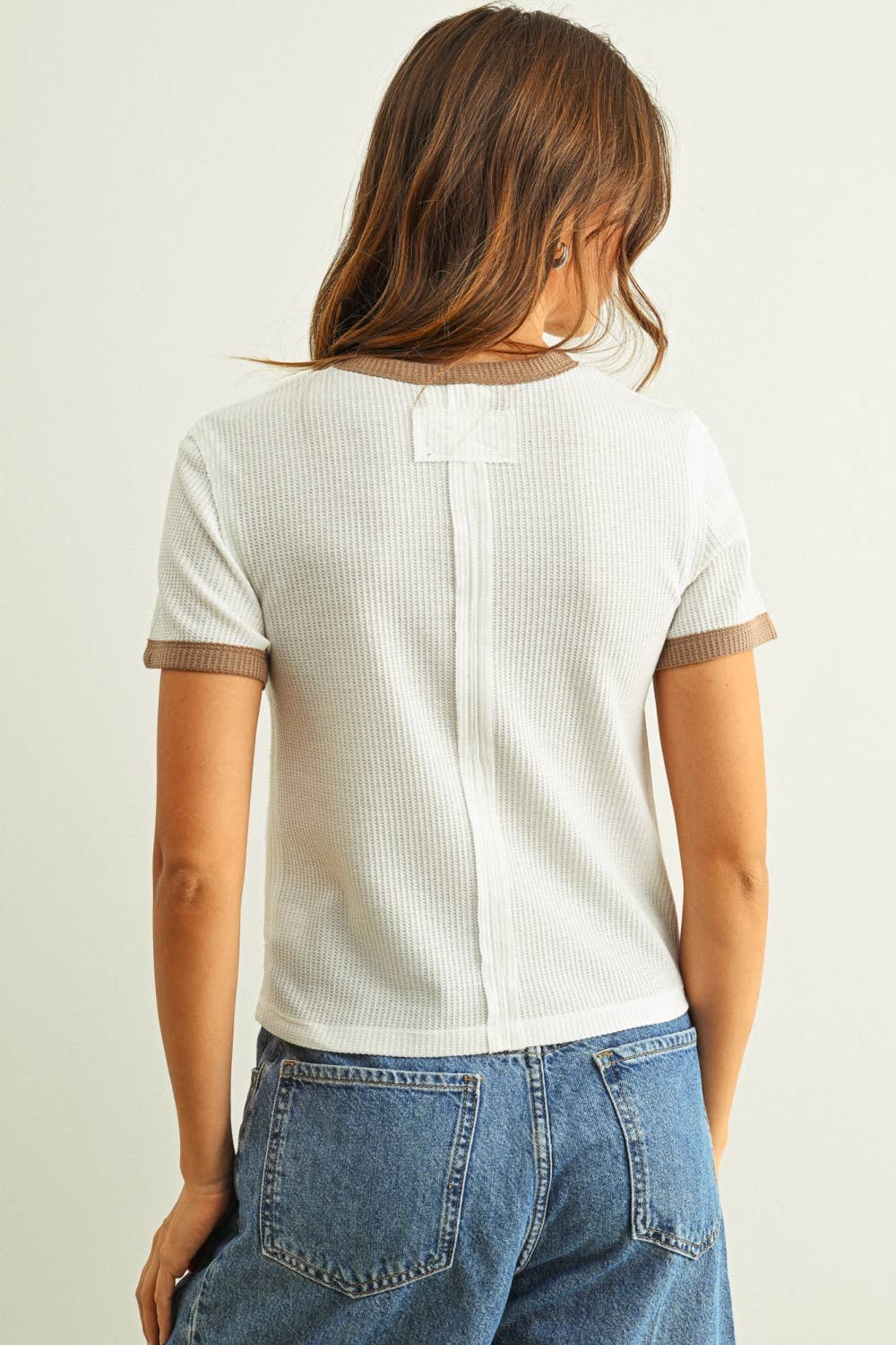 Woman wearing a white ribbed knit top with brown trim and blue jeans on a plain background