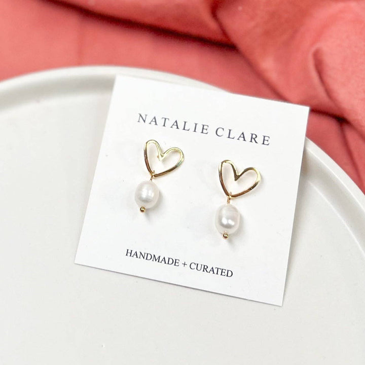 A pair of Natalie Clare handmade and curated drop earrings featuring gold heart-shaped wire frames and white pearls, displayed on a white card over a pink fabric background.