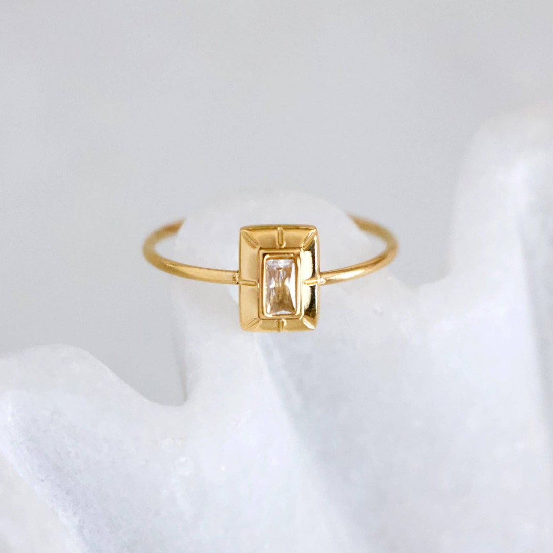 A gold ring with a rectangular bezel-set clear gemstone, displayed on a white textured stand against a soft light gray background.