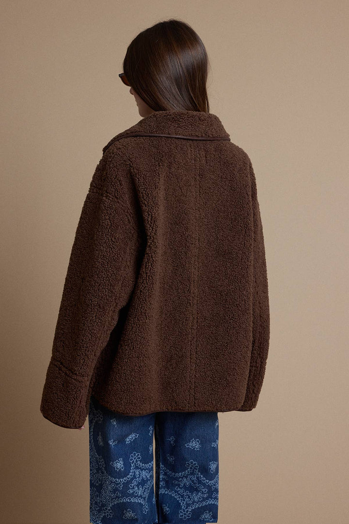 A woman shown from the back wearing an oversized brown teddy fleece jacket and blue paisley-print pants against a plain beige studio background.