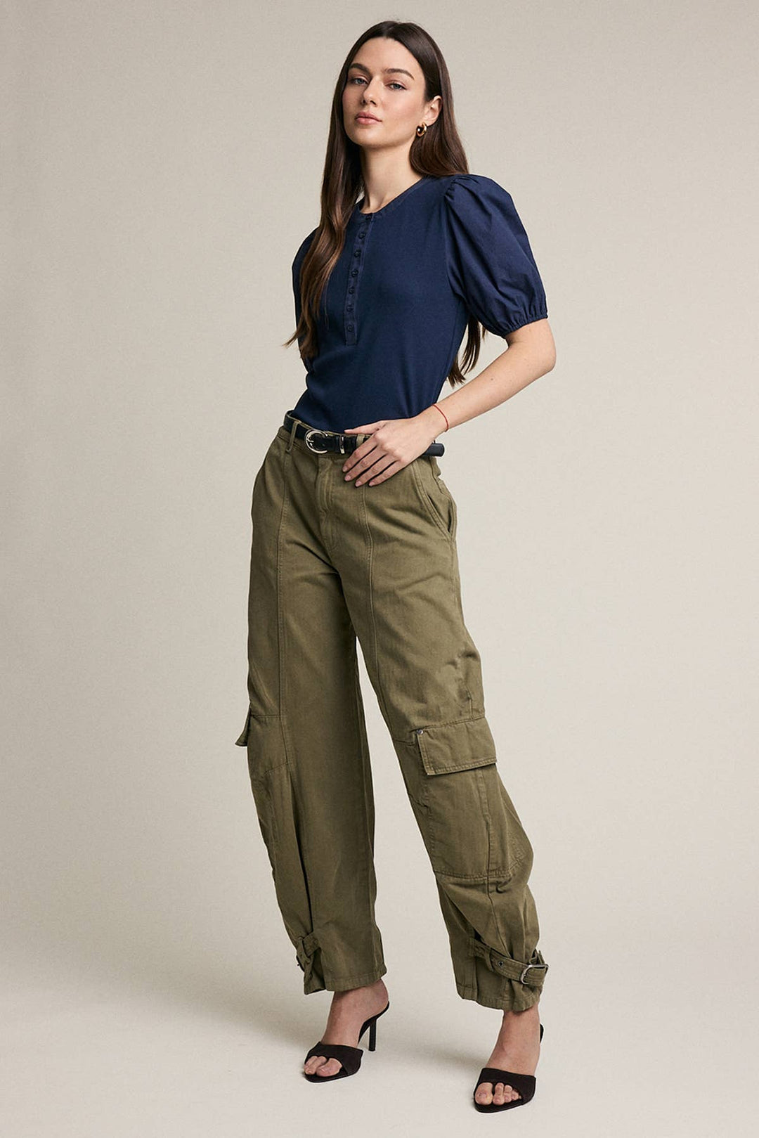 A woman modeling a navy blue puff-sleeve blouse and olive green cargo pants with a black belt, paired with black heeled sandals, against a solid light beige studio background.