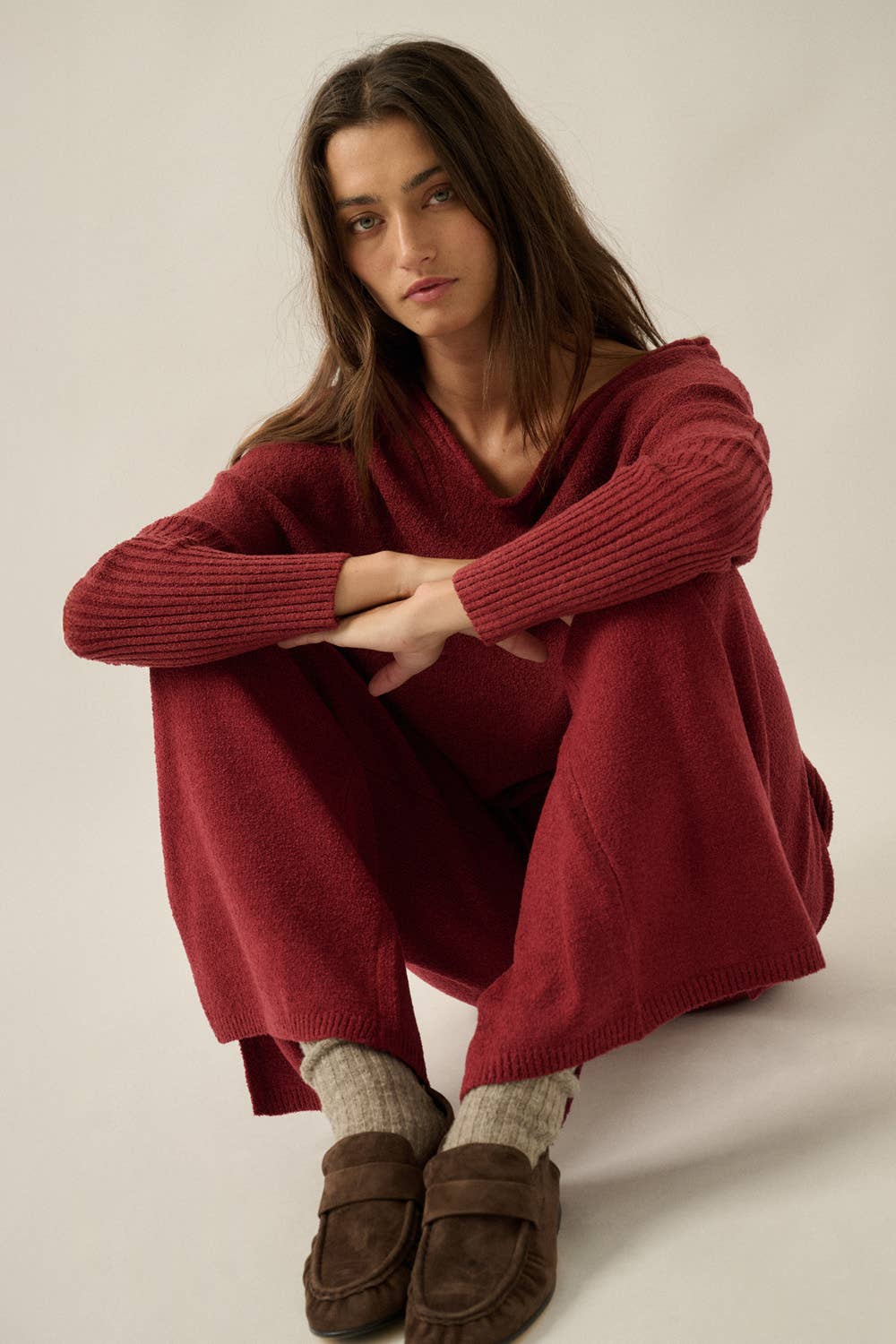 A woman crouches in a studio wearing a loose, oversized ribbed knit sweater dress in deep burgundy red, paired with beige socks and brown suede loafers, against a plain light gray background.