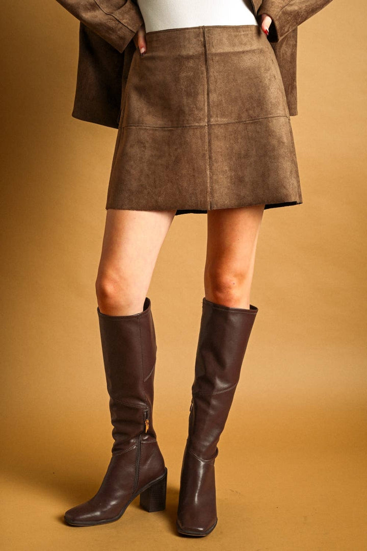 A model wearing a brown suede-like mini skirt, a matching brown jacket, a white top, and dark brown knee-high leather boots, standing against a solid warm brown background.