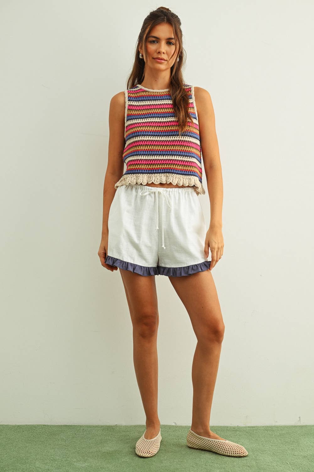 Woman wearing a colorful striped sleeveless crochet knit top and white shorts on a plain background