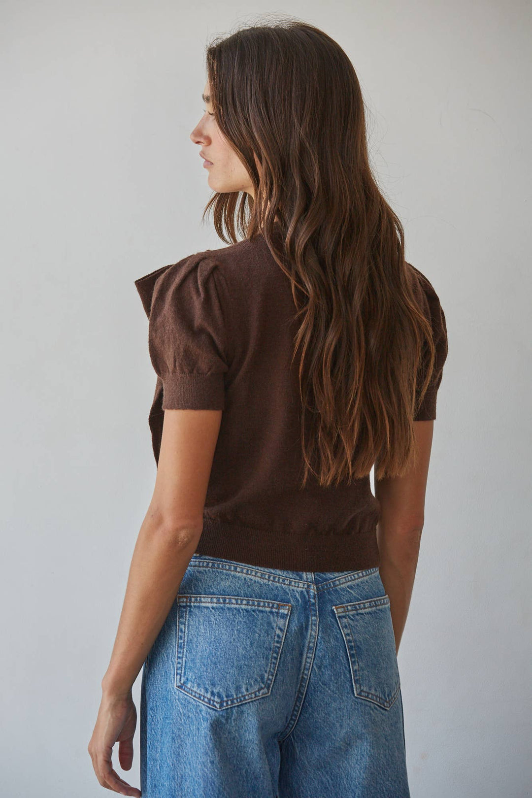 A woman with long brown hair is shown from the back wearing a dark brown short-sleeve puff-sleeve knit top and blue jeans against a plain light gray background.