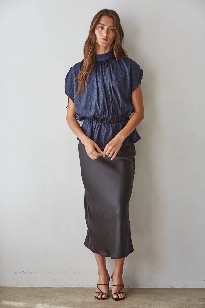 A woman stands against a plain light gray wall wearing a dark blue satin sleeveless peplum top and a dark charcoal gray midi skirt, paired with brown strappy sandals.