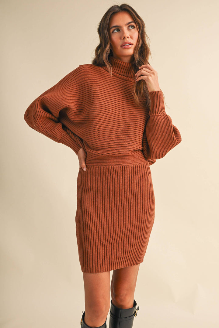 A woman modeling a rust-orange ribbed knit mock-neck sweater dress with balloon sleeves, paired with black ankle boots, against a solid beige studio background.
