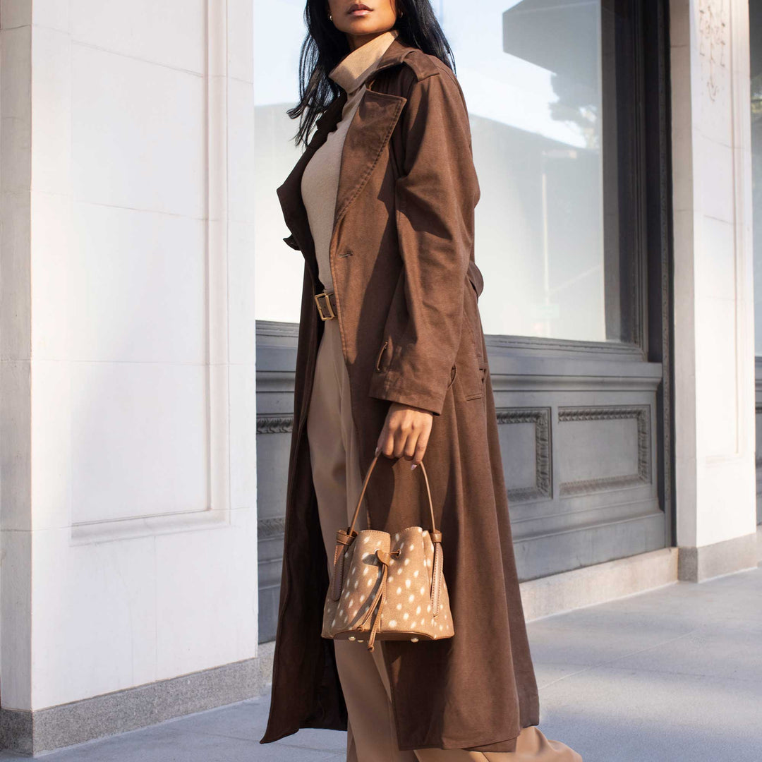 A woman wearing a long brown trench coat over a beige top and brown pants, holding a beige polka-dot bucket bag, standing in front of a modern building with white columns and a large window.