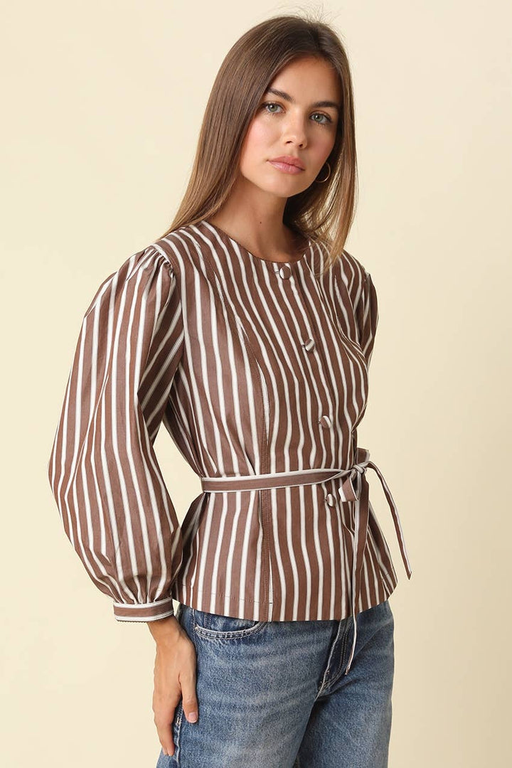 A woman wearing a brown and white vertically striped long-sleeve blouse with a tie waist belt, paired with blue jeans, against a solid light beige background.