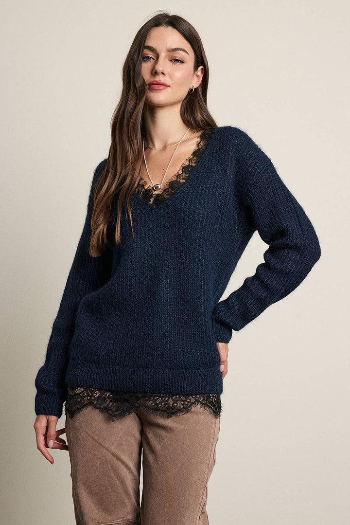 A woman modeling a navy blue ribbed knit sweater with black lace trim along the V-neck and hem, paired with brown pants, against a plain beige studio background.
