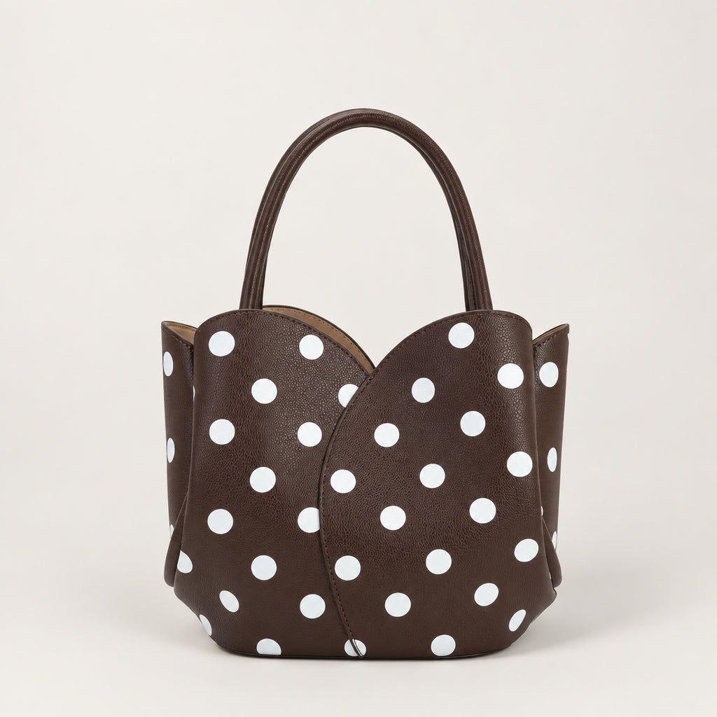 A dark brown vegan leather handbag with a white polka dot pattern, featuring a structured shape, double top handles, and a small black recycling symbol on the upper left corner.