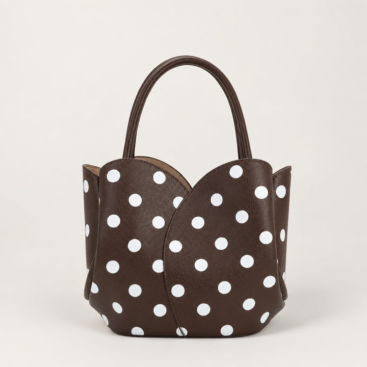 A dark brown vegan leather handbag with a white polka dot pattern, featuring a structured shape, double top handles, and a small black recycling symbol on the upper left corner.
