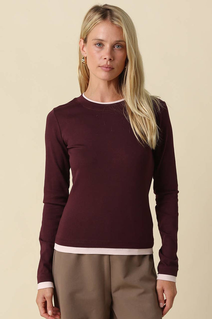 A blonde woman modeling a long-sleeve crewneck sweater in deep burgundy with contrasting light pink trim at the collar, cuffs, and hem, paired with taupe trousers against a solid beige background.