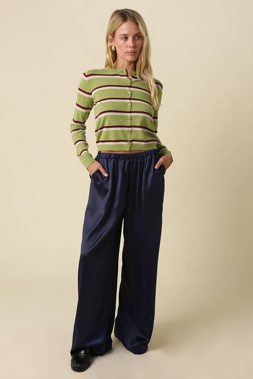 A woman modeling a green and burgundy striped long-sleeve button-up cardigan with dark navy wide-leg pants, standing against a solid beige studio background.