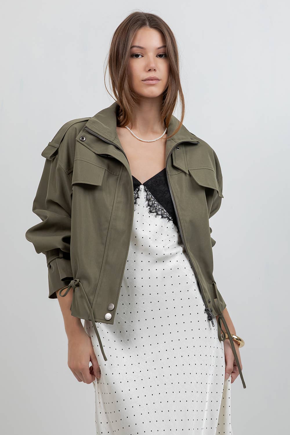 A woman models an olive green utility jacket over a white polka dot dress with a black lace insert, set against a plain light gray studio background.