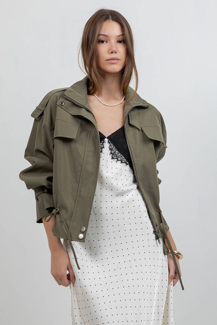A woman models an olive green utility jacket over a white polka dot dress with a black lace insert, set against a plain light gray studio background.