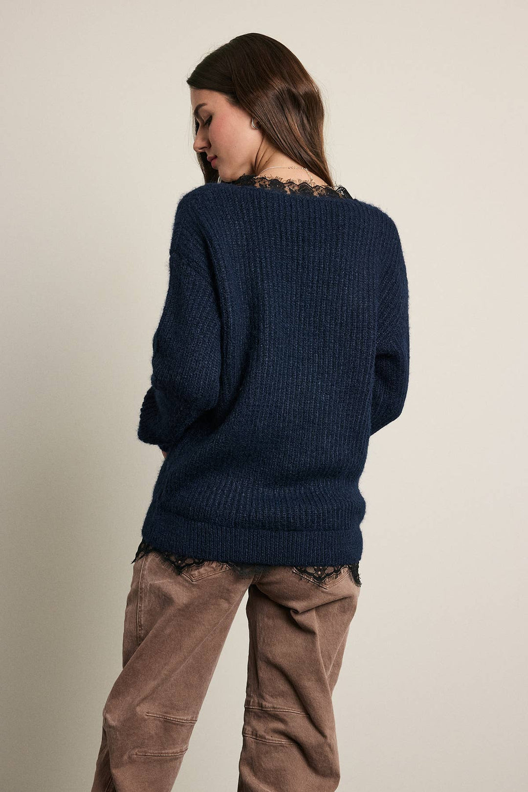A woman shown from the back wearing a dark navy blue ribbed knit sweater with black lace trim at the neckline and hem, paired with brown pants, against a plain light beige background.