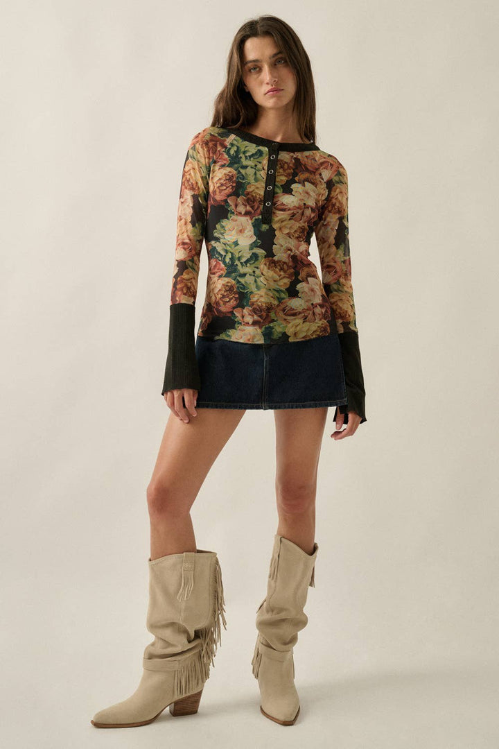 A model wearing a long-sleeve floral knit top with black cuffs, a dark denim mini skirt, and beige fringed boots, standing against a plain beige studio background.