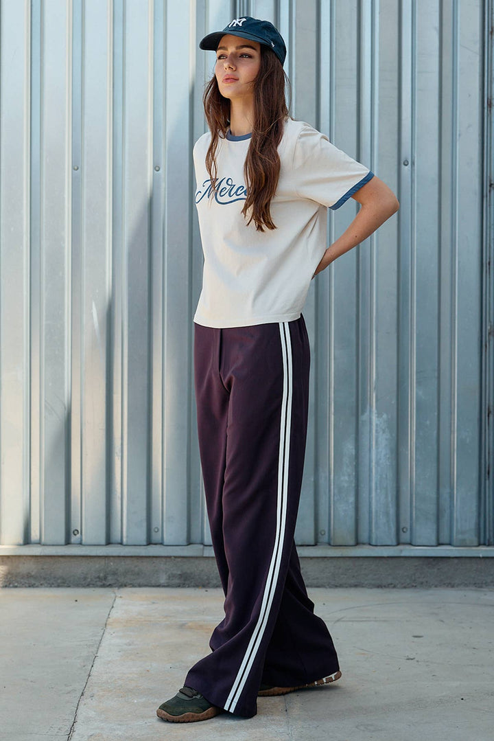 A woman stands outdoors against a corrugated metal wall wearing a cream graphic T-shirt with blue trim, burgundy colored wide-leg track pants with white side stripes, and a dark baseball cap.