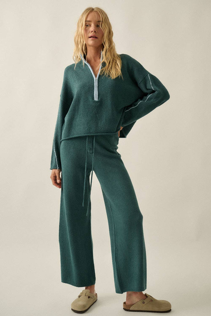 A woman modeling a matching two-piece knit loungewear set in a deep teal green color, featuring a half-zip long-sleeve top and wide-leg pants with a drawstring waist, styled with beige slip-on shoes against a plain beige studio background.
