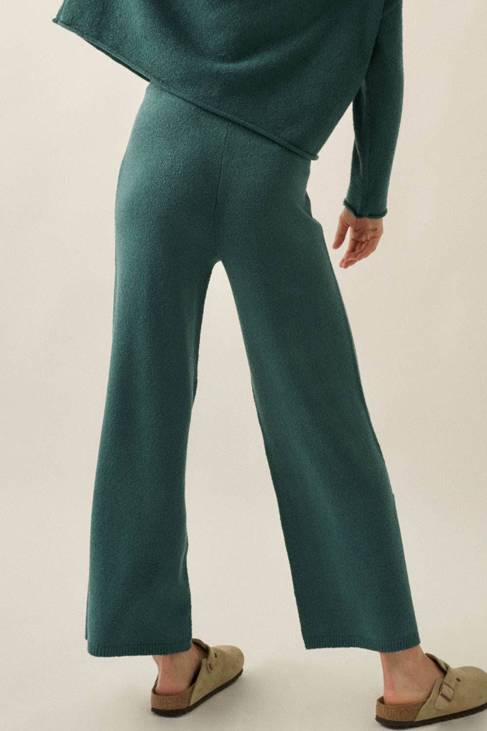 A woman wearing a matching two-piece knit outfit in a deep teal green, featuring wide-leg cropped pants and a draped long-sleeve top, styled with tan slip-on mule sandals against a plain light beige studio background.